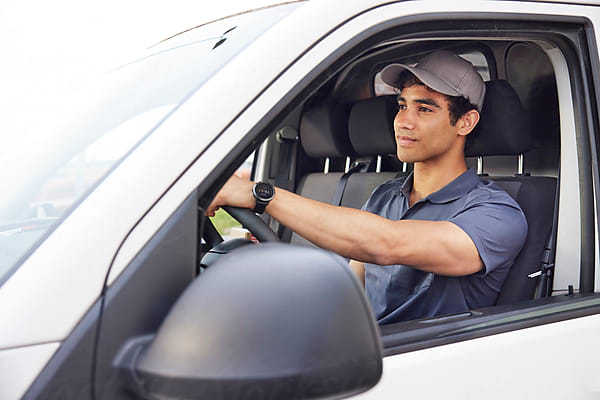 Young Hispanic male courier in uniform and cap looking away and steering white van while working on sunny summer day in city.